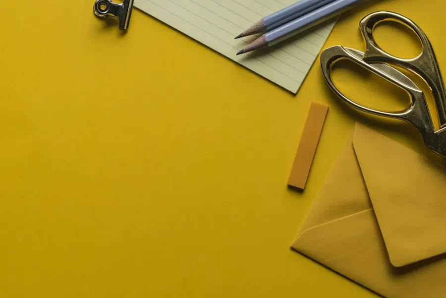 Scissors, pencils, lined paper and an envelope on a bright yellow desk.